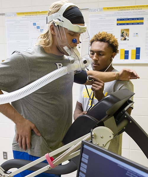 Male student measuring the pulse and oxygen level as another student runs on treadmill. 